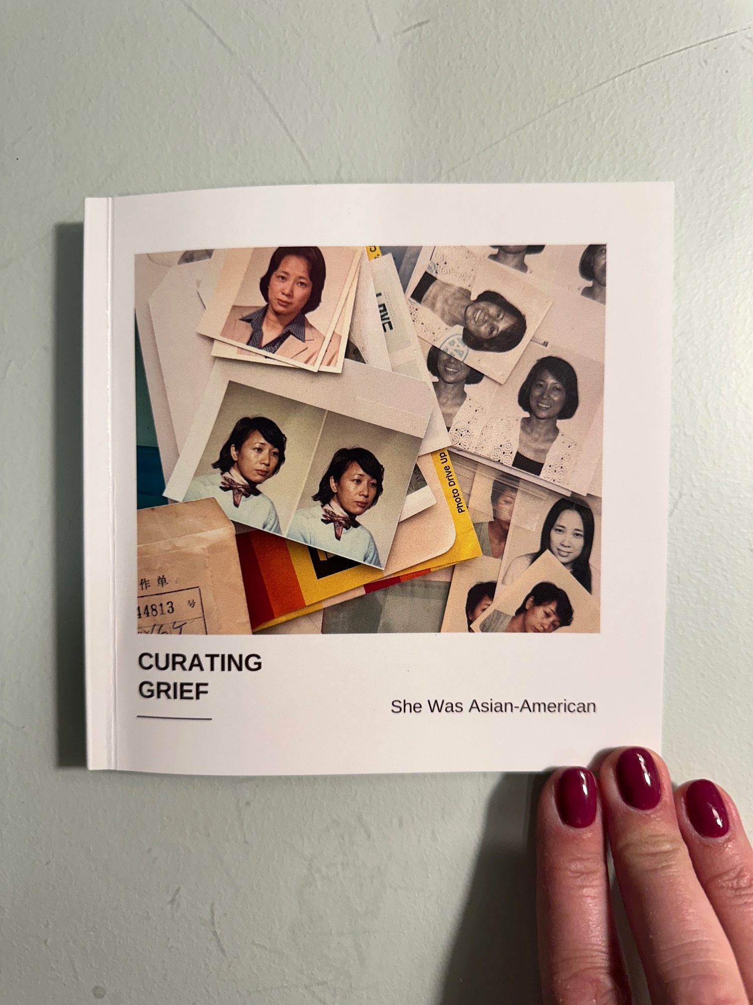 Small square book titled Curating Grief: She Was Asian-American with picture of scattered passport photos of an Asian woman at different ages. A hand with red nail polish is resting on the book.